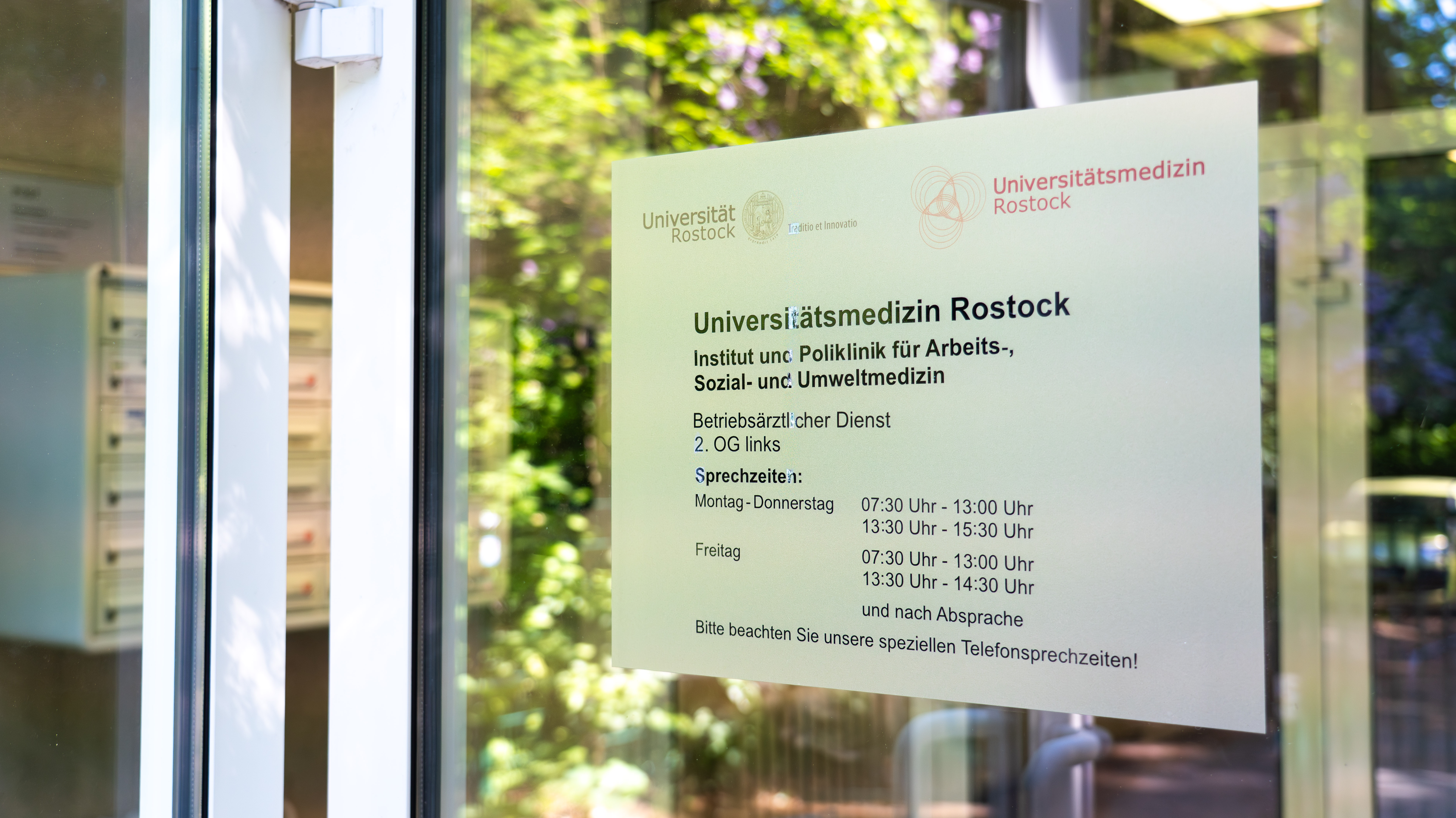 An der Glastür eines Gebäudes hängt ein Schild mit Informationen zur Universitätsmedizin Rostock, Institut und Poliklinik für Arbeits-, Sozial- und Umweltmedizin, Öffnungszeiten und Kontakthinweisen.