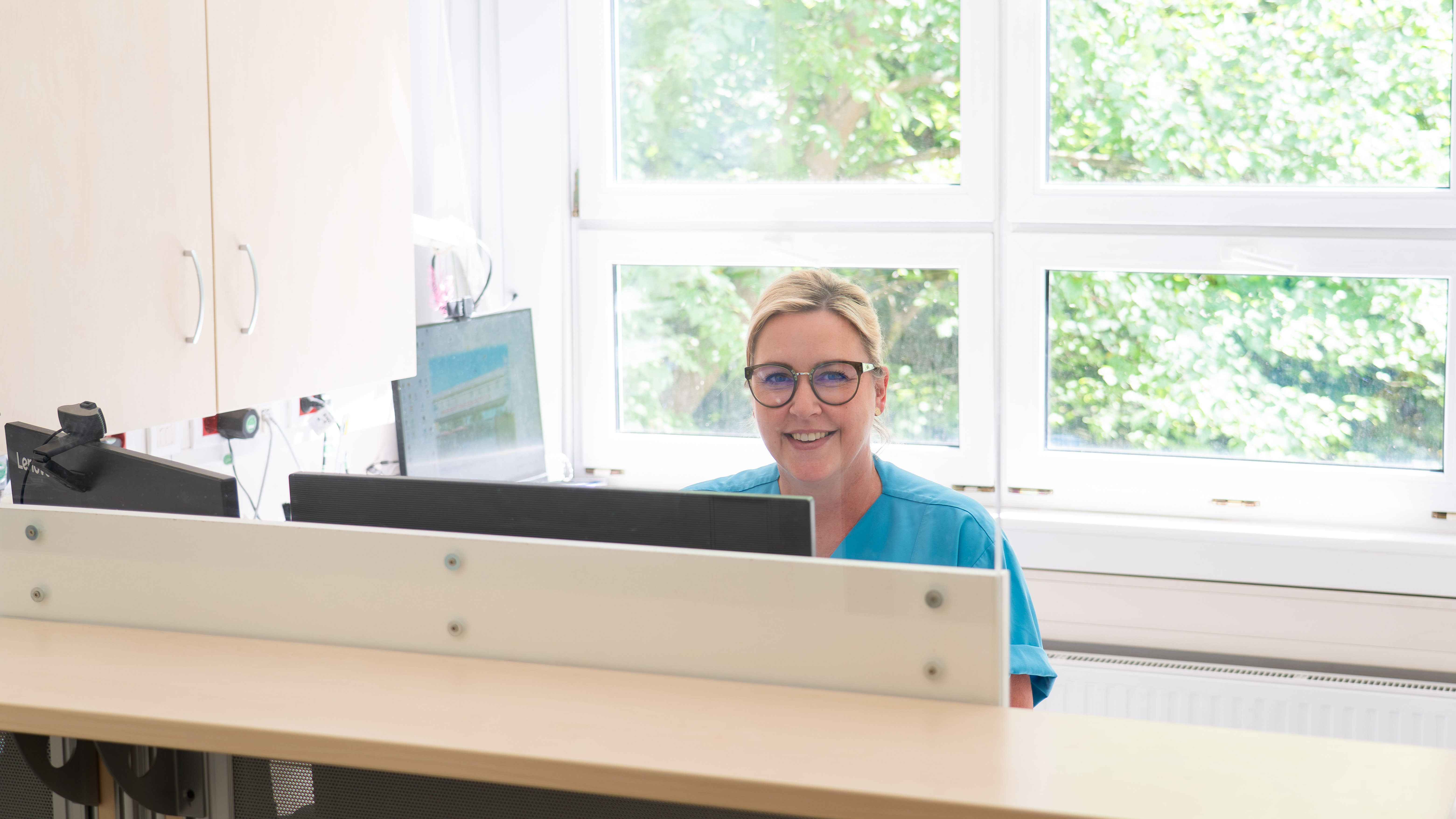 Frau mit Brille und blauem Oberteil sitzt lächelnd hinter einem Schreibtisch mit Computer in einem hellen Büro mit großen Fenstern