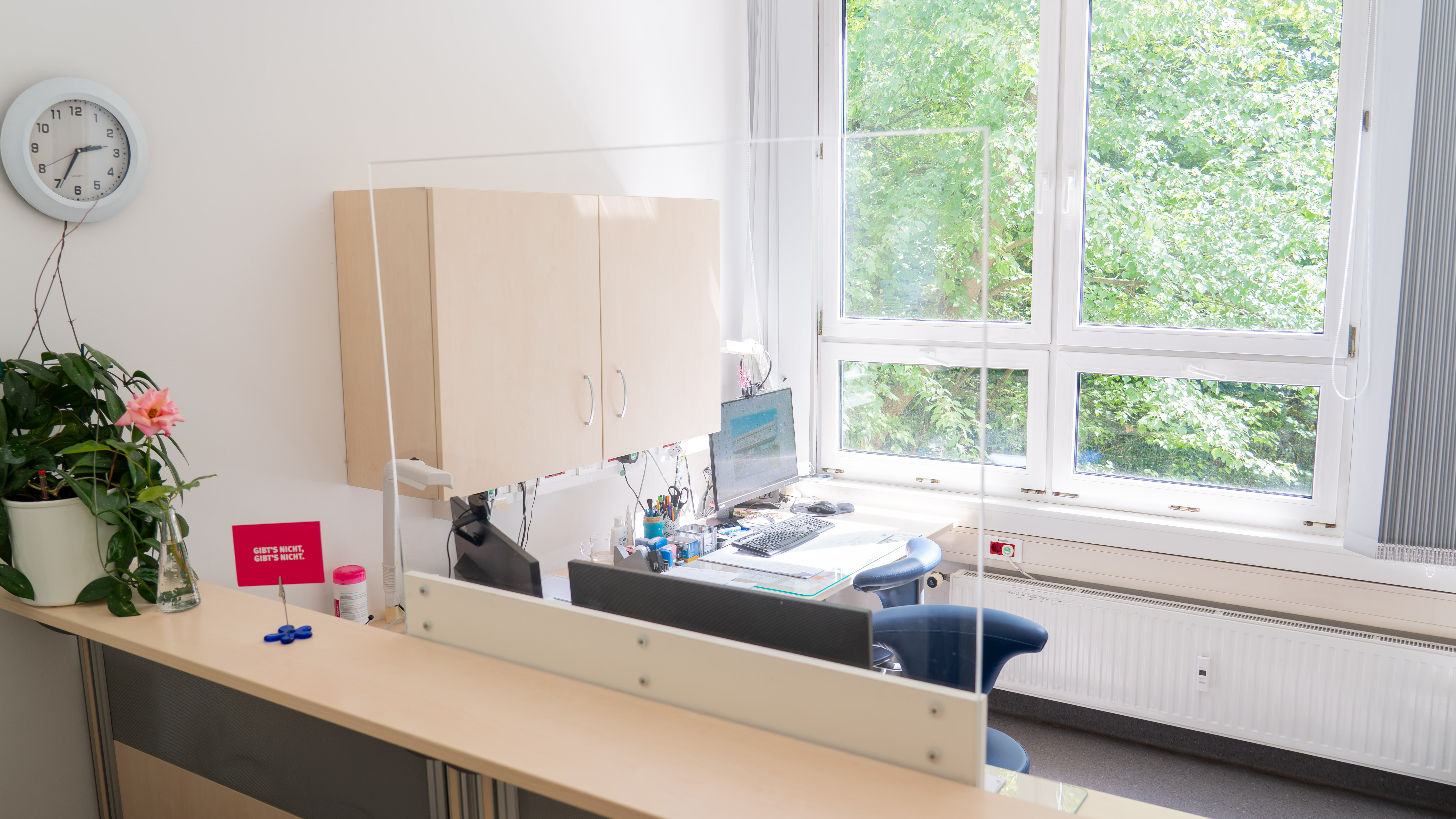 Heller Büroraum mit Schreibtisch, Computer, Schrank, Uhr an der Wand und großen Fenstern mit Blick auf grüne Bäume