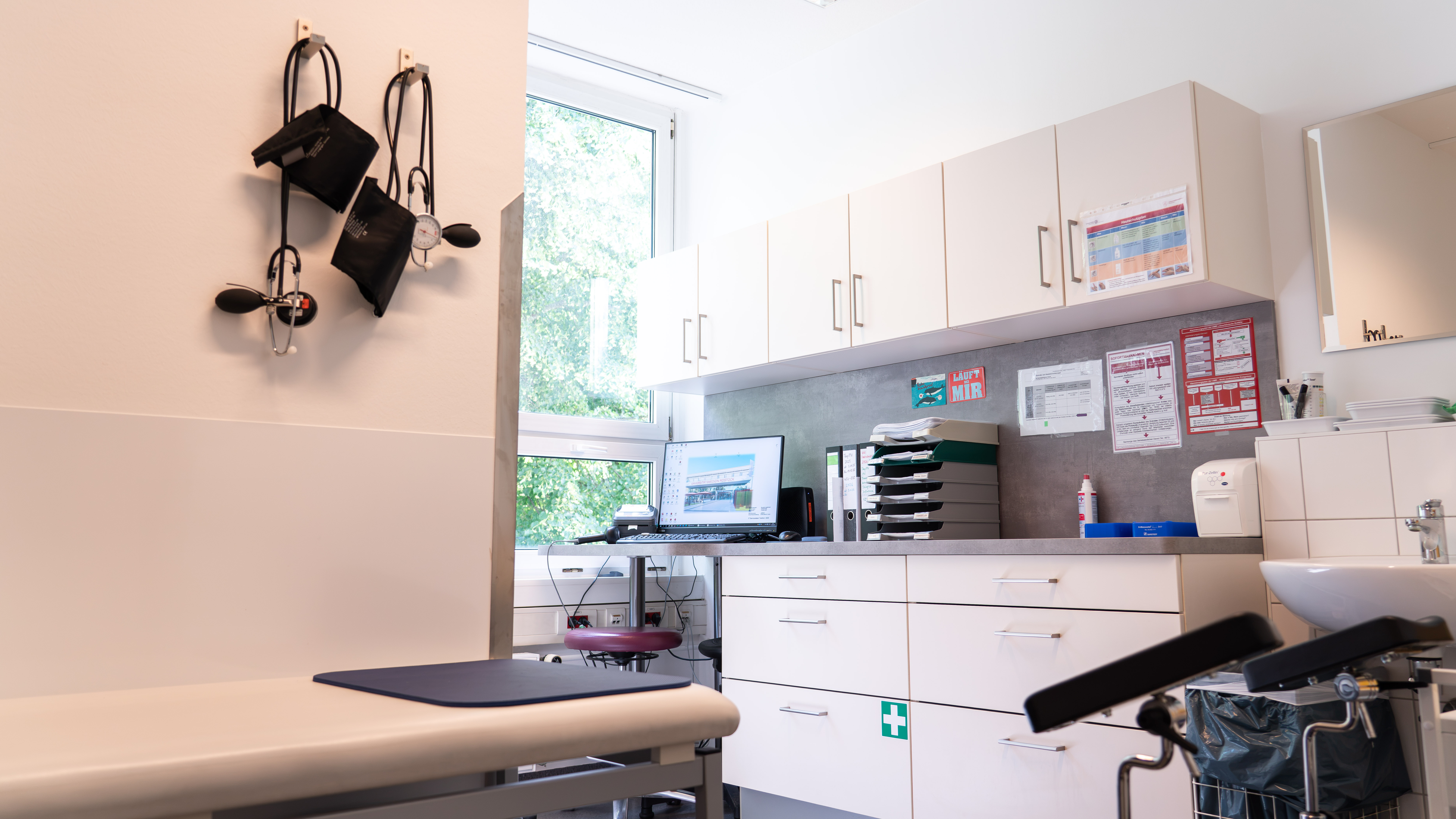 Behandlungszimmer mit Untersuchungsliege, Hocke, Computer auf der Arbeitsfläche und medizinischen Utensilien an der Wand und auf der Arbeitsfläche