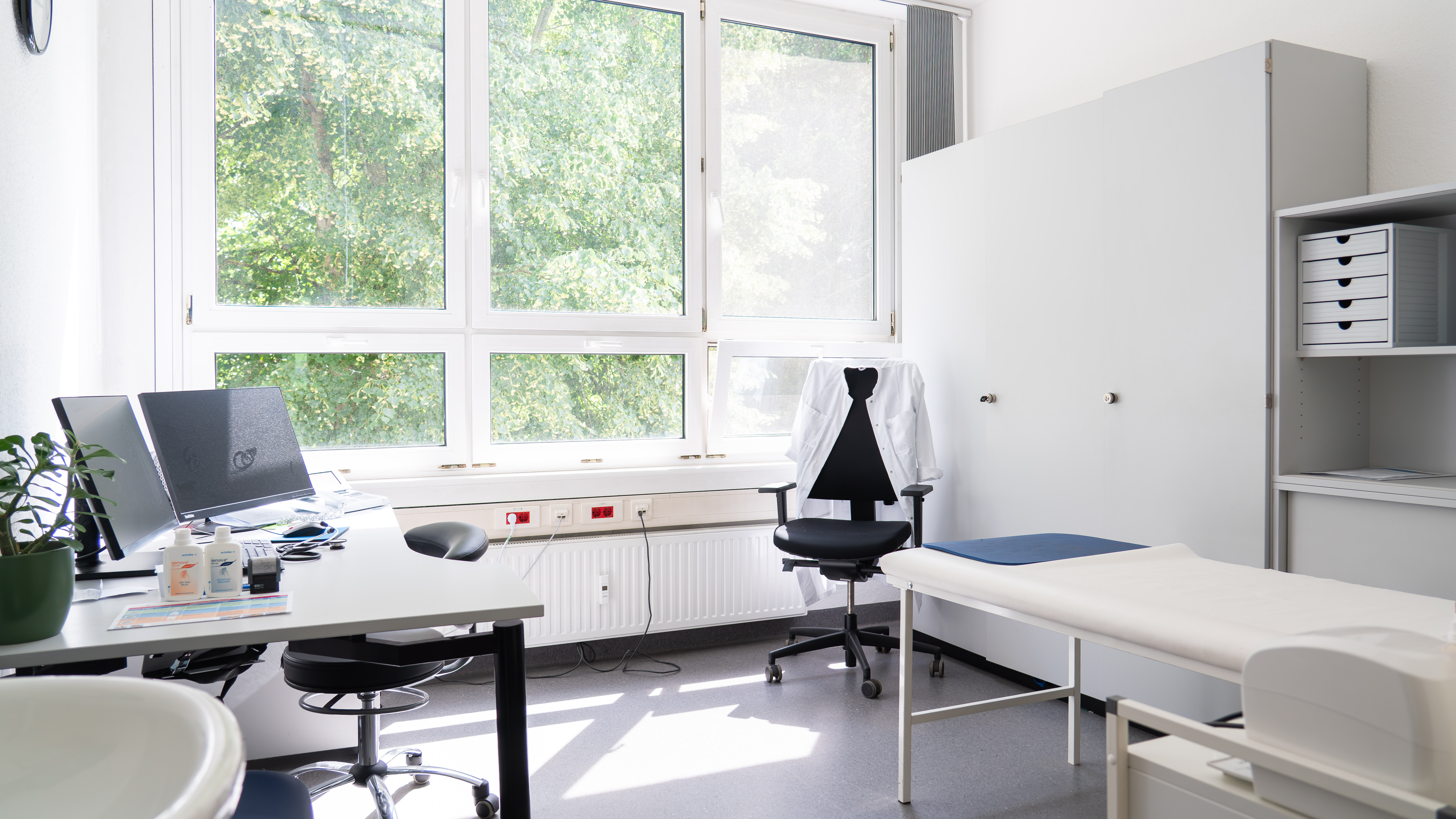Helle Arztpraxis mit Untersuchungsliege, Schreibtisch mit Computer, zwei Stühlen und großem Fenster mit Blick auf Bäume