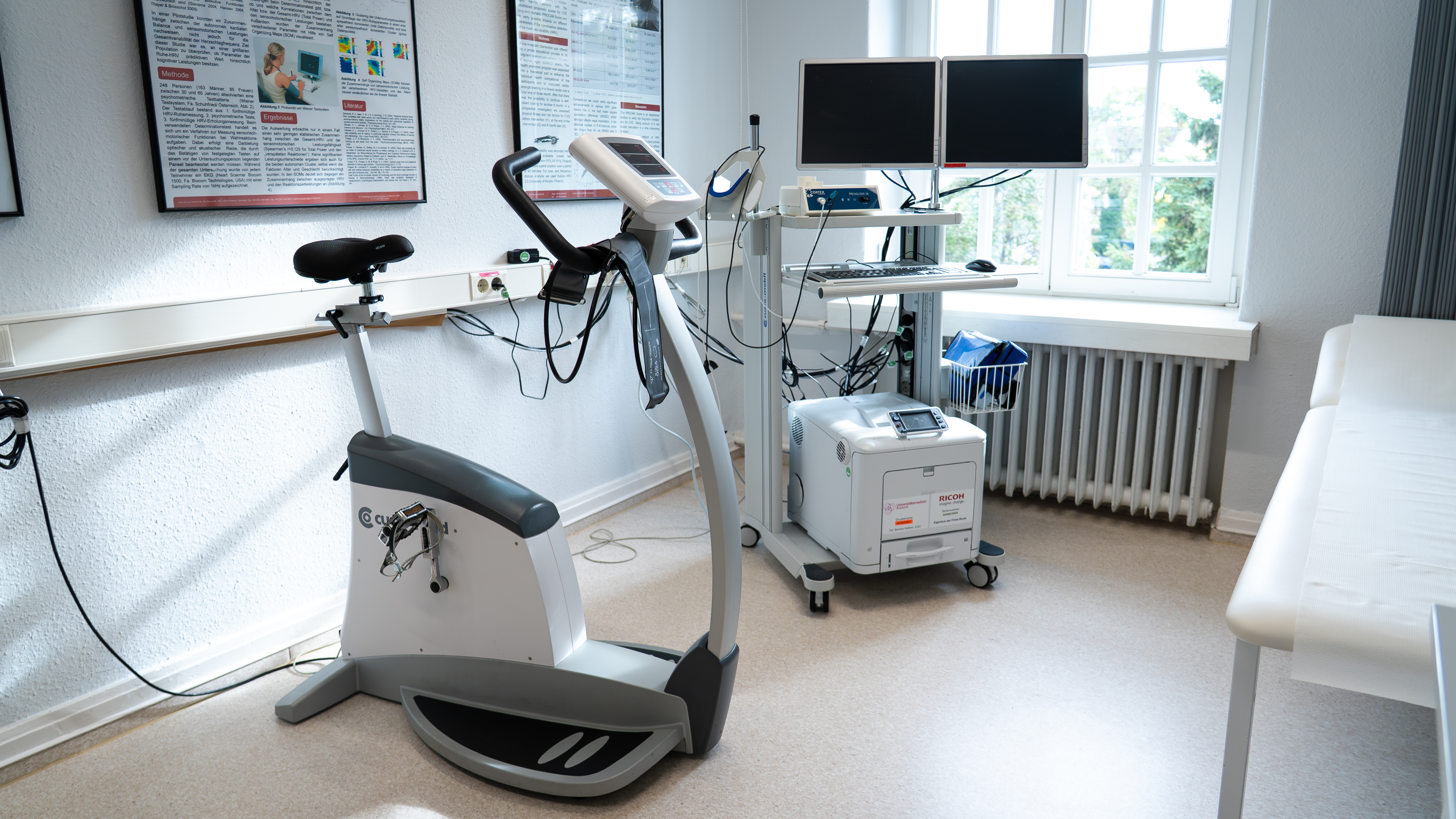 Stationäres Ergometerfahrrad neben medizinischem Gerät mit Monitoren in hellem Untersuchungsraum