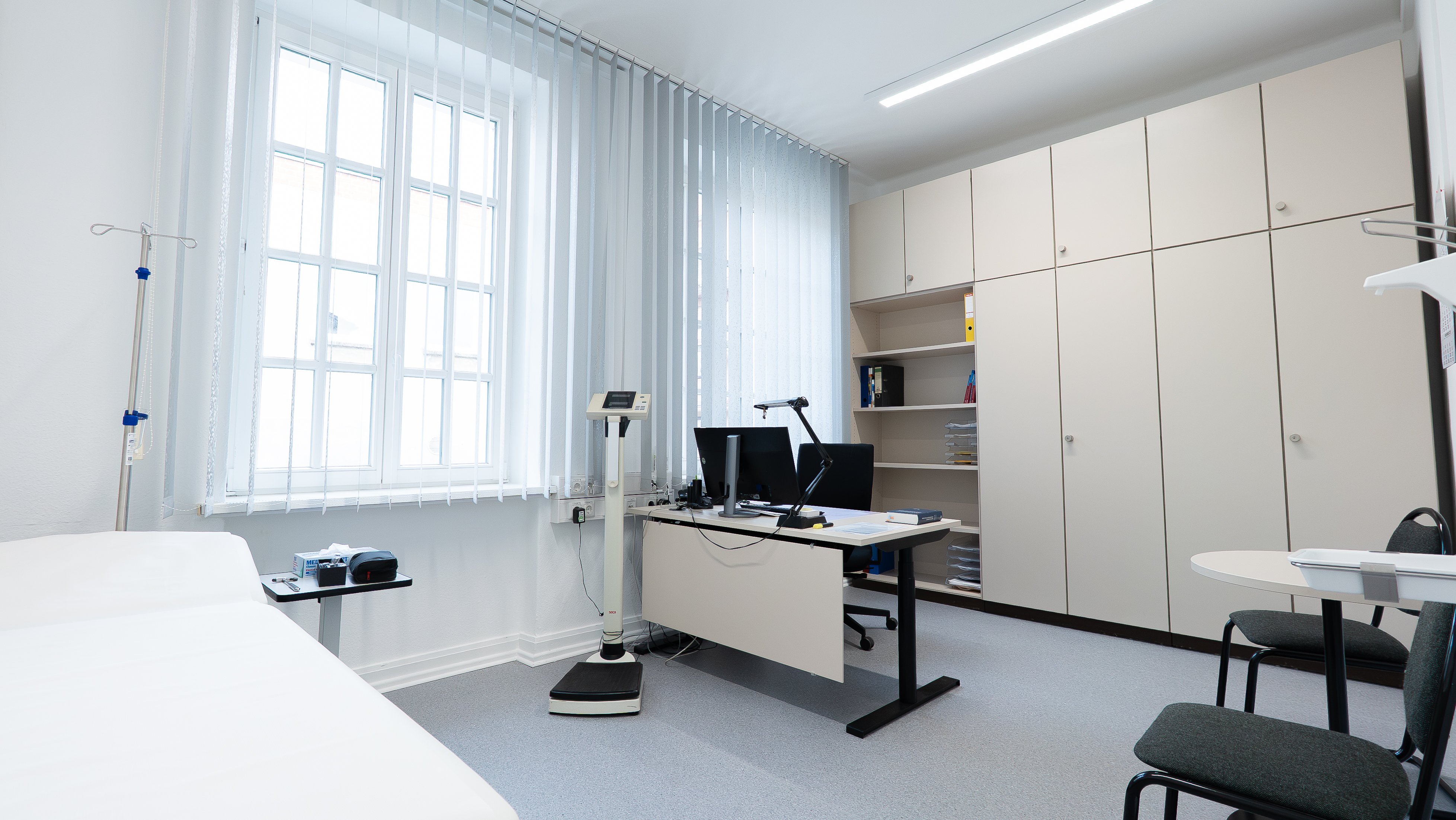 Helles Arztzimmer mit Untersuchungsliege, Infusionsständer, Waage, Schreibtisch mit zwei Monitoren und Stuhl sowie großem Schrank mit offenen Fächern